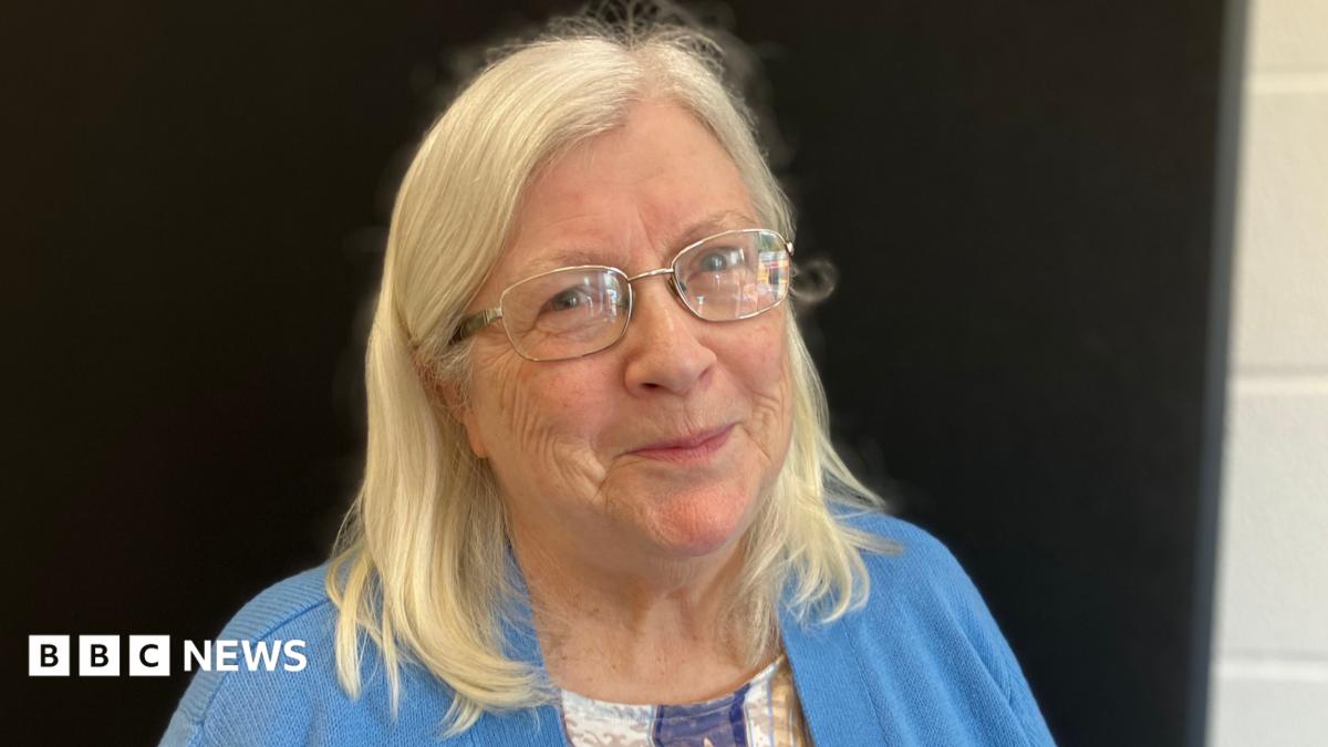 A woman in glasses with shoulder length blonde hair. She is wearing a blue cardigan and is stood against a black background