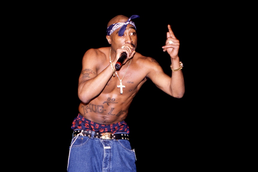 A Black man, Tupac Shakur, with no shirt, chain with crucifix around neck, tattoos, briefs showing, bandana, holding mic