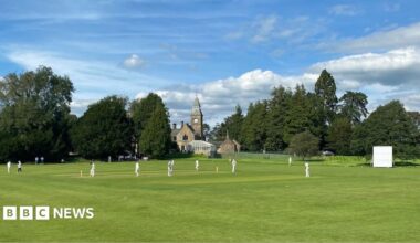 Sedgwick Cricket Club members playing cricket on their pitch in Sedgwick. It is a large grassy patch of land with a clock tower and church in the background. The players are all wearing white.