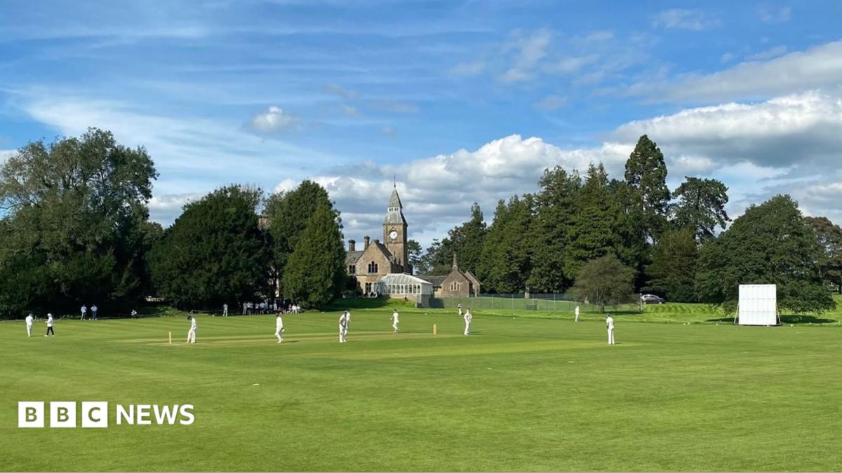 Sedgwick Cricket Club members playing cricket on their pitch in Sedgwick. It is a large grassy patch of land with a clock tower and church in the background. The players are all wearing white.