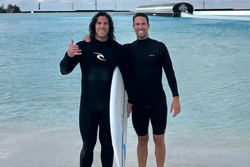 Two men in wetsuits stand together near water