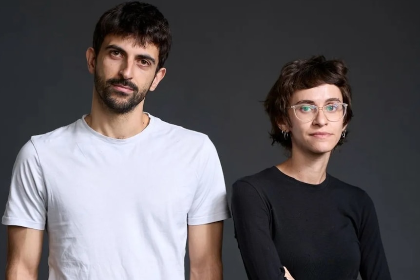 A man wearing a white t-shirt stands next to a woman with glasses wearing a black t-shirt.