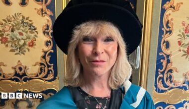 Toyah Willcox is smiling at the camera. She has shoulder-length blonde hair and is wearing a blue ceremonial gown and a black Tudor bonnet with a flowery top underneath.
