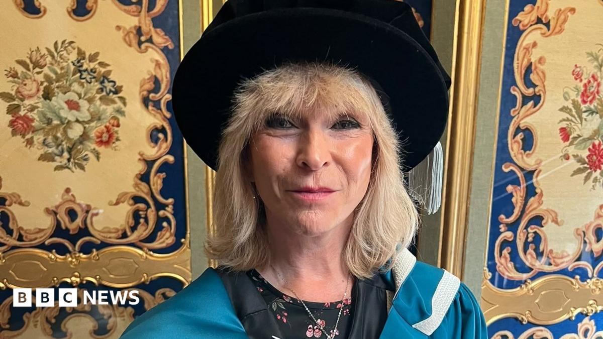 Toyah Willcox is smiling at the camera. She has shoulder-length blonde hair and is wearing a blue ceremonial gown and a black Tudor bonnet with a flowery top underneath.