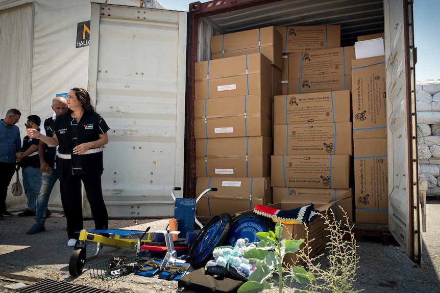 A shipping container full of large cardboard boxes, with a wheelchair and knitted rugs out the front illustrating what's inside.