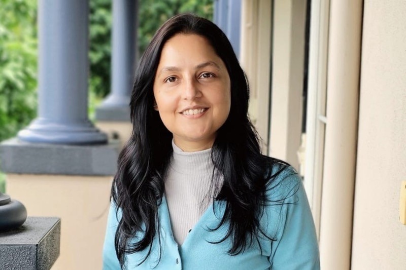 A woman with long dark hair in a grey skivy and light blue cardigan smiling at camera, standing outside