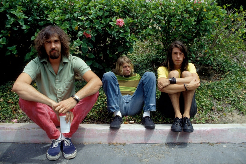 Three young musicians sitting on a ledge outside , arms wrapped around legs