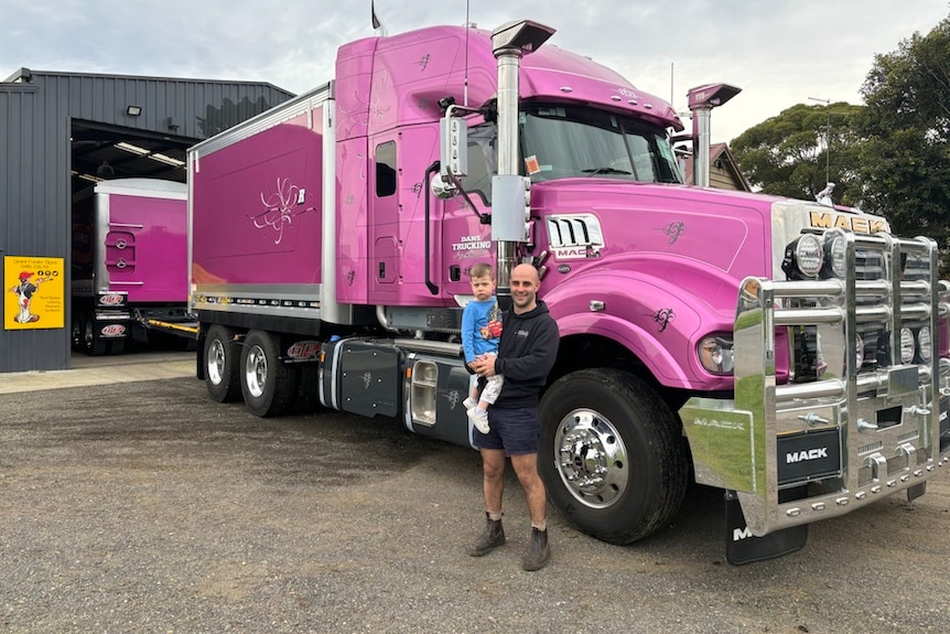 A man holding a young child stands in front of a pink semi trailer