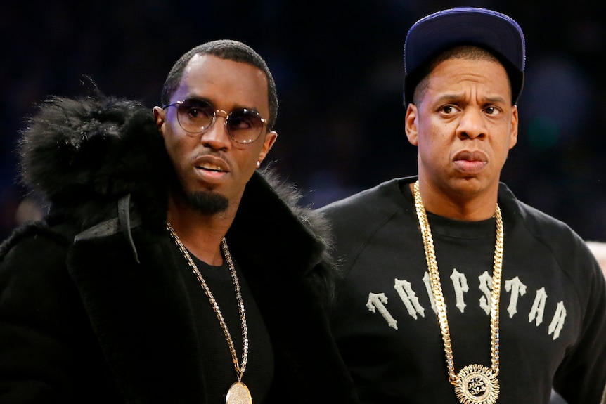 Two men wearing black clothes and big gold chains look sideways, one has a concerned expression