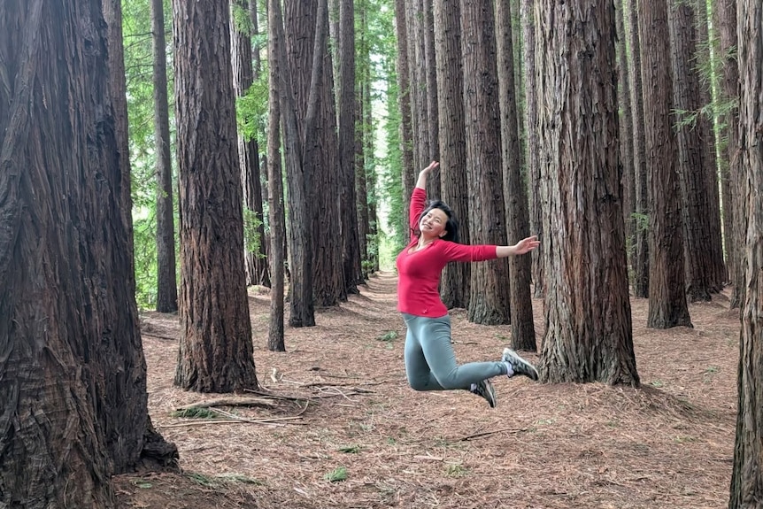 Woman wearing red long-sleeved top and jeans jumps high amid a line of large trees.