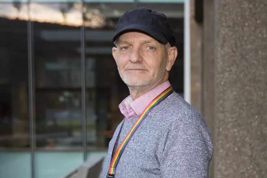 A man stands a looks into the camera, wearing a hat.