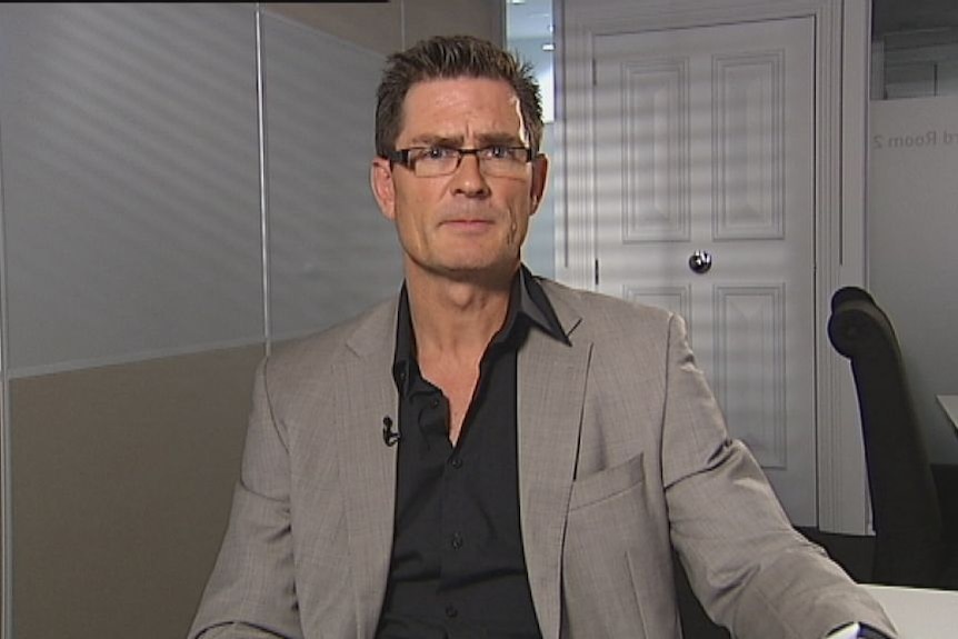 Marcus Padley wears glasses and a suit and looks at the camera with a serious expression.