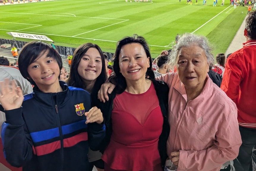A mother and her boy and girl stand with their grandmother at a football match. Field and crowd is behind them.