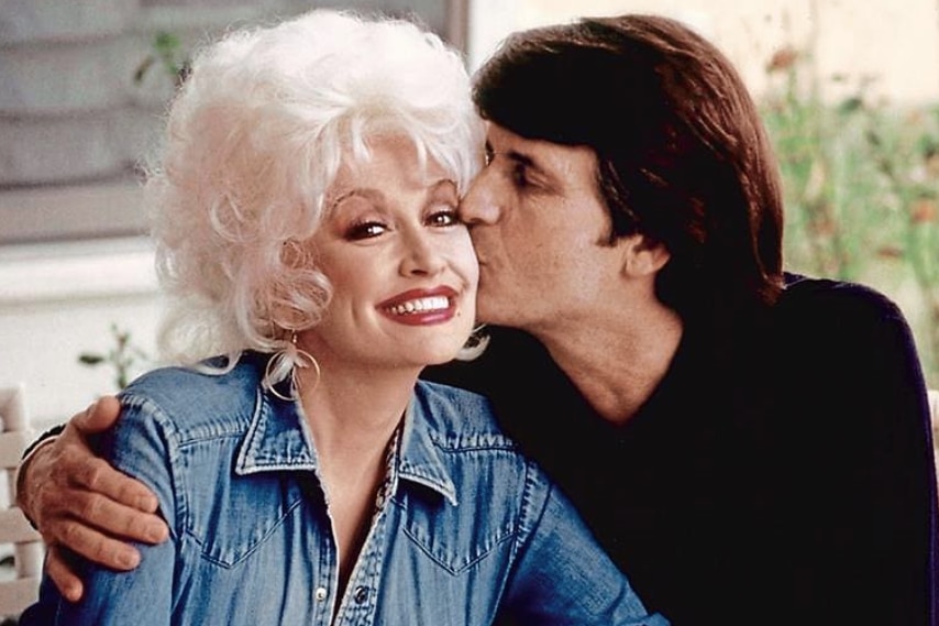 An older woman with blonde voluminous hair smiles while a man seated next to her kisses her cheek.