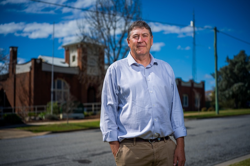 A man looking serious in a blue shirt stands across the road of the council building which is in background