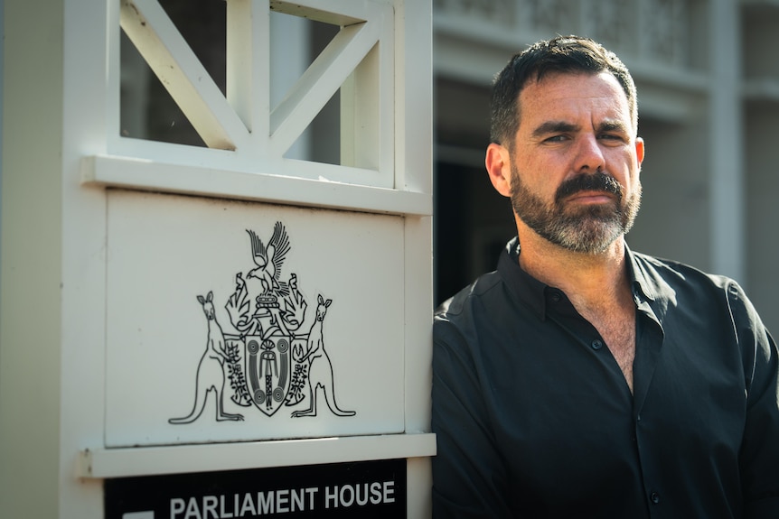 Chansey Paech has dark brown short-cut hair and a brown, graying beard, standing near an NT parliament sign, serious expression.