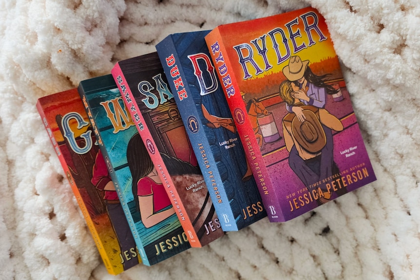 Five books from the same cowboy romance series stacked on each other asthetically over a plush blanket.