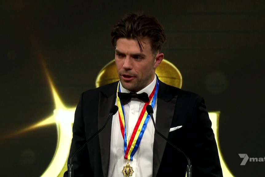 A man dressed in a tuxedo speaks at a lectern wearing a gold medal with red, yellow and blue ribbon.