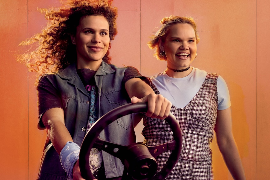 Ruby Henaway and Lila McGuire, two young Indigenous women, pose smiling, Henaway holding a steering wheel.