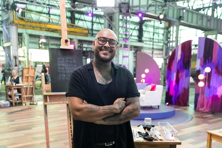 A man with brown skin stands smiling at the camera with his arms folded in front of him. An easel and art supplies behind him