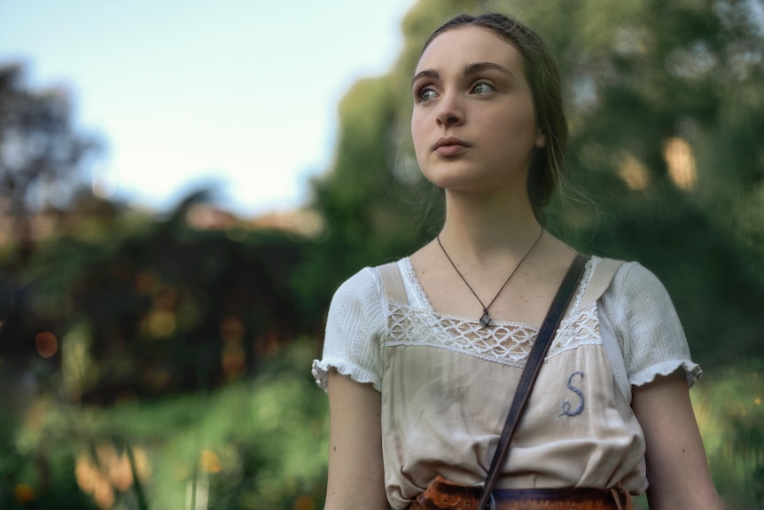 A TV still of Chloe Jean Lourdes, a teenage girl, with a placid expression, looking slightly up, and in an old-fashioned dress.