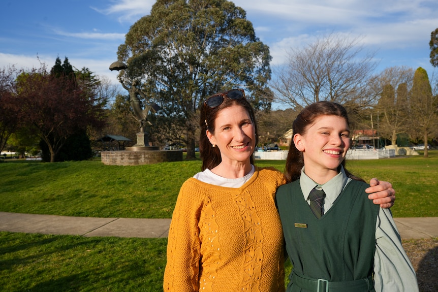 A woman and her daughter, both with long dark hair and smiling, stand in a park.