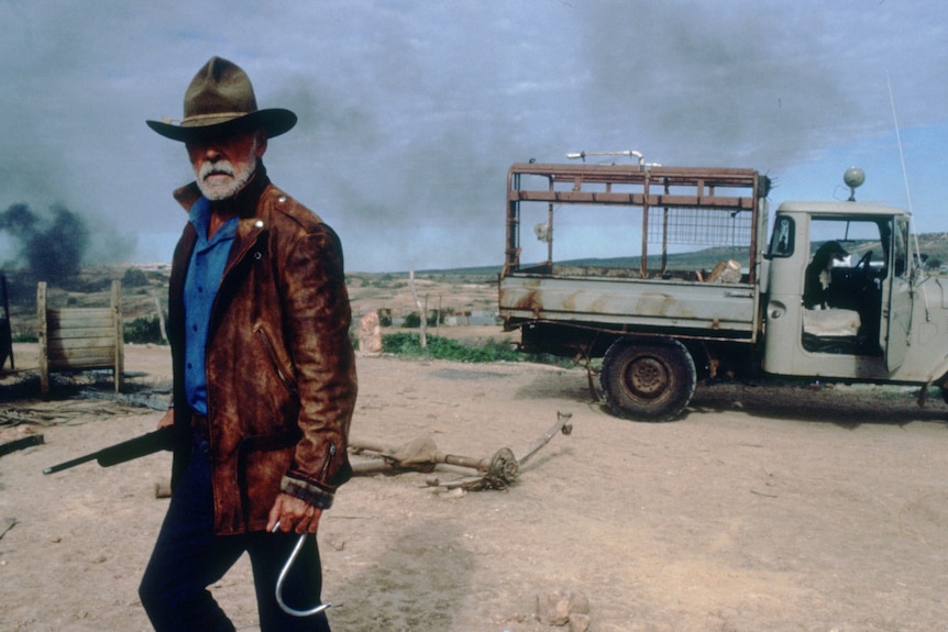 A man wearing a cowboy hat standing out in the open, dirt road, truck behind him