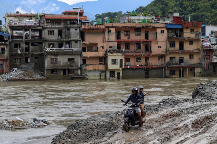A motorcycle, with two people aboard, atop a muddy outcrop, above a river of brown water, in front of destroyed buildings.