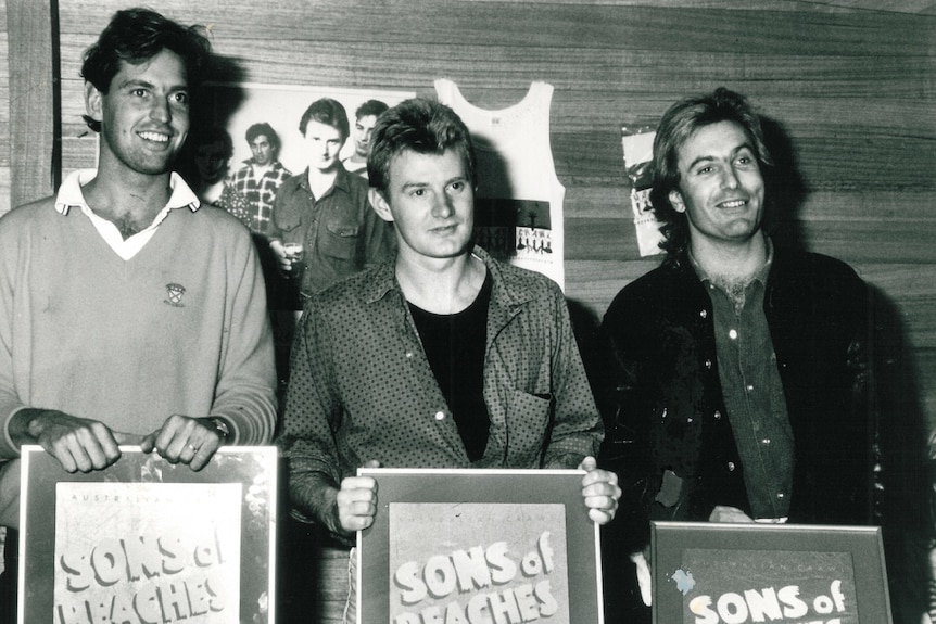 Australian Crawl members hold Sons of Beaches award plaques against a wood-paneled wall