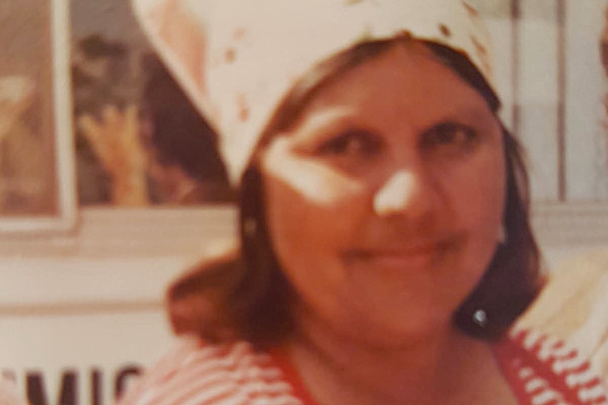 A young woman, with a bandana scarf on her head, smiles shyly at the camera.