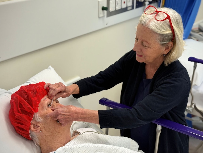 A woman with short blonde hair puts drops in the eyes of a patient lying on a bed with a red cap on her head.