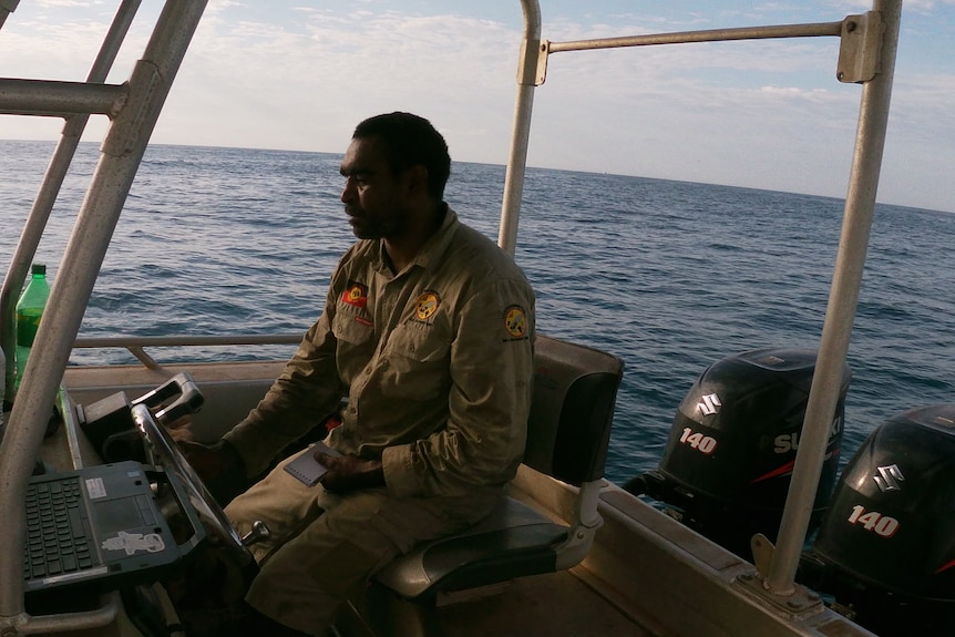 A man in a khaki shirt steers a boat through blue waters