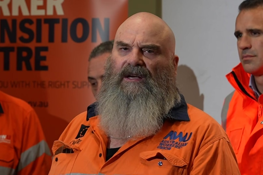 A man with a beard is standing before a row of people wearing orange work tops.
