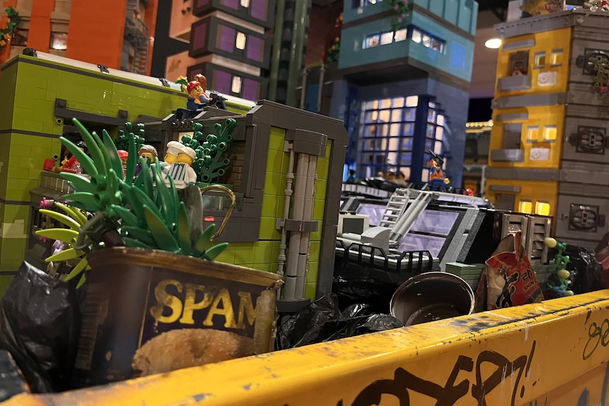A yellow skip bin with graffiti is full of rubbish, but lego people are living amongst it.