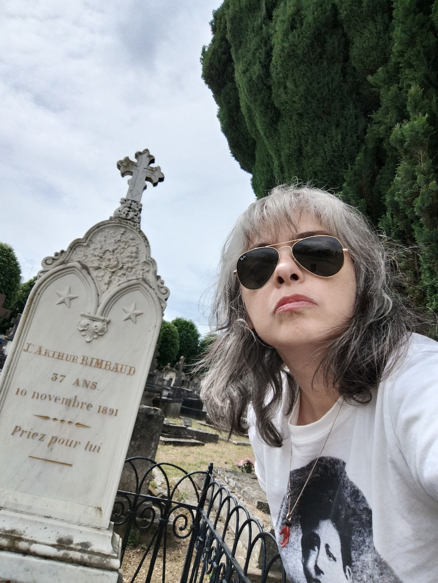 A selfie photo of a woman with shoulder length grey and black hair wearing glasses with a headstone in a cemetery