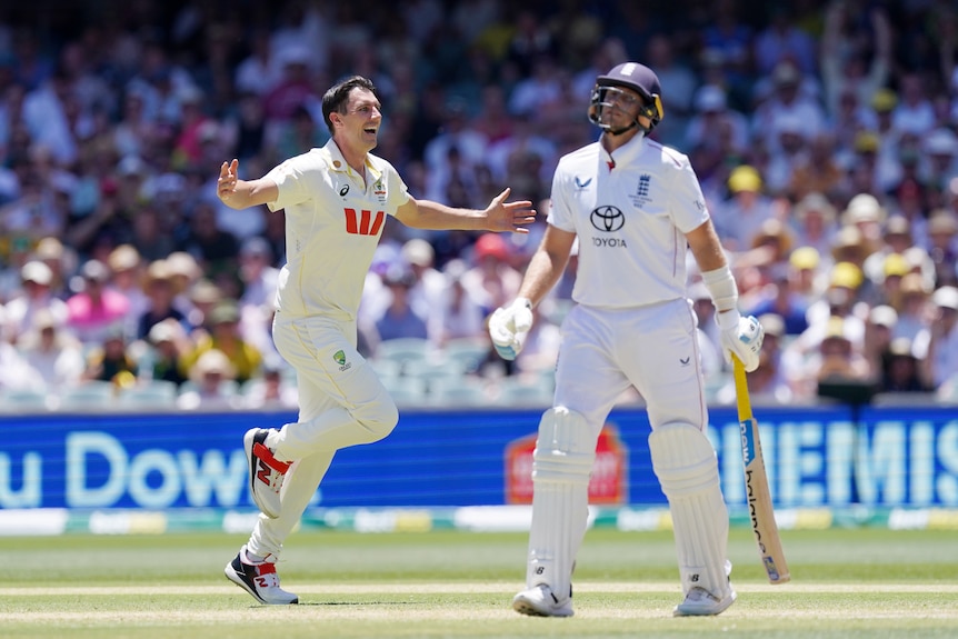 A cricketer with black hair runs and smiles with arms outstretched as another cricketer looks glum next to him.