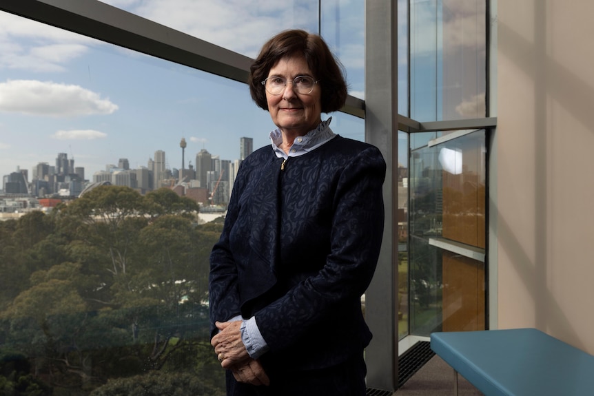 lady in twin set stands before window with Sydney skyline behind her