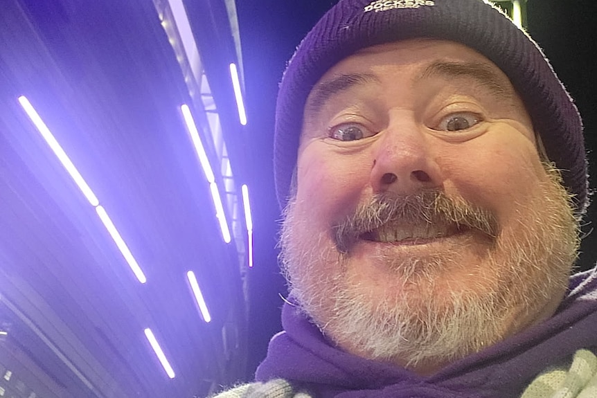 A smiling, middle-aged man with a grey beard wears a beanie and scarf.