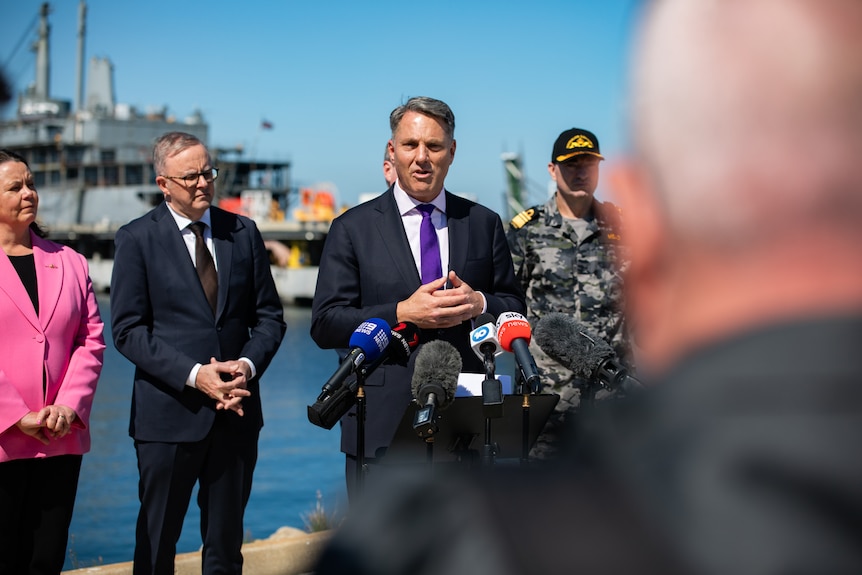 Richard Marles, in a suit, stands in front people at a harbour.