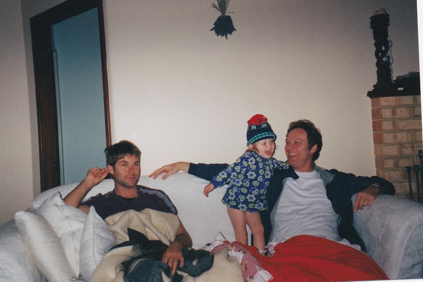 Old photo of a man in his mid-30s sat on a white couch with a female toddler and a man in his early 40s.