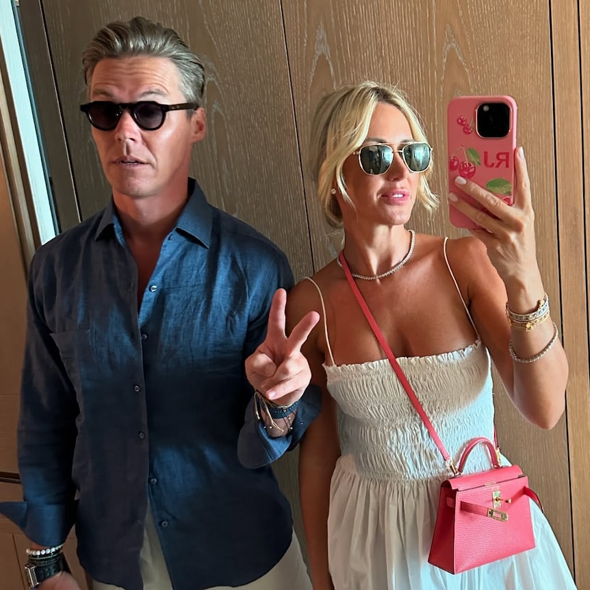 A man with sunglasses and his hair slicked back gives a peace sign next to his wife.