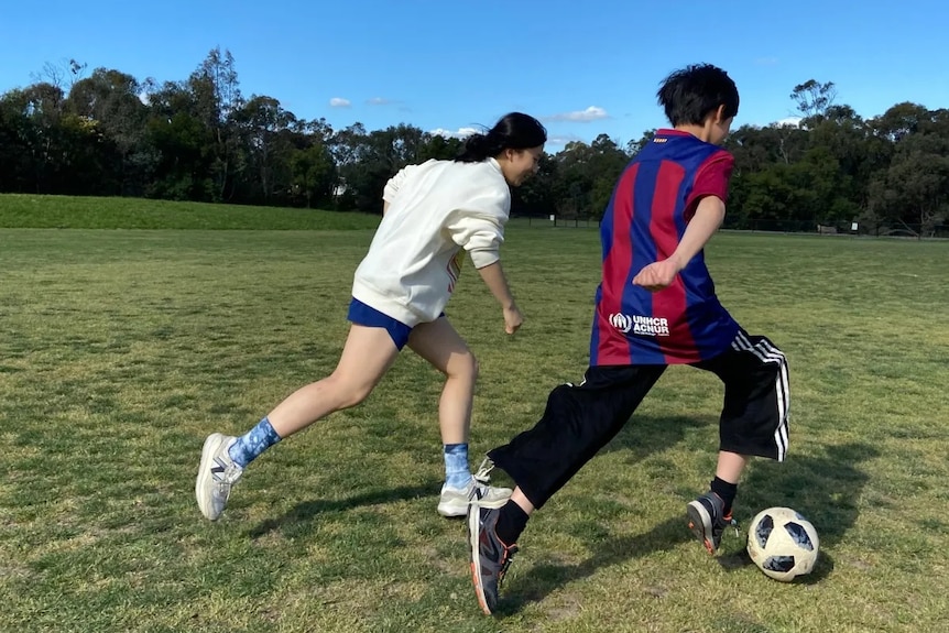 A boy and a girl with their backs turned run on a grass field on a sunny day playing soccer.