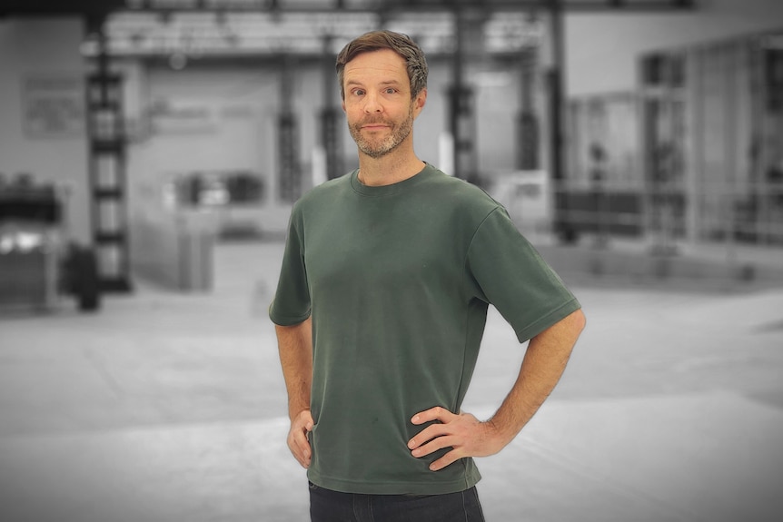 Man wearing dark green t-shirt standing with arms on hips in the middle of a grey warehouse