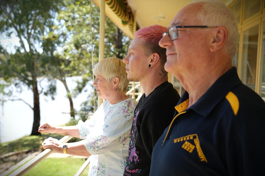 Three people stand on a balcony looking out over a river from the first floor of a house.