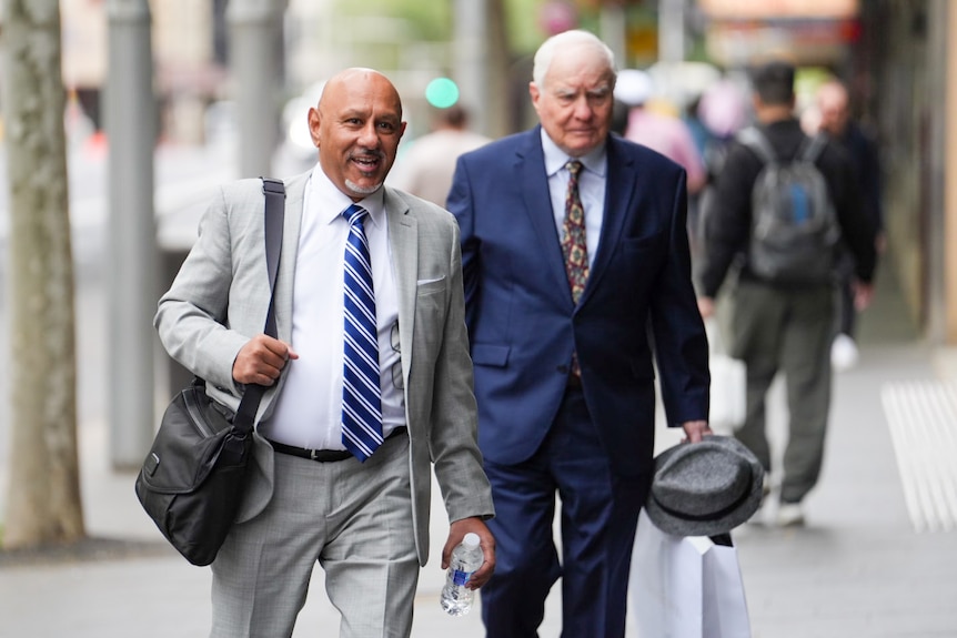 Shane has tan-coloured skin, is bald and walks carrying a satchel.