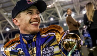 Robert Lambert is grinning and sticking his tongue out. He is wearing a black baseball hat and his black, gold and blue racing outfit. He is cradling his gold trophy.
