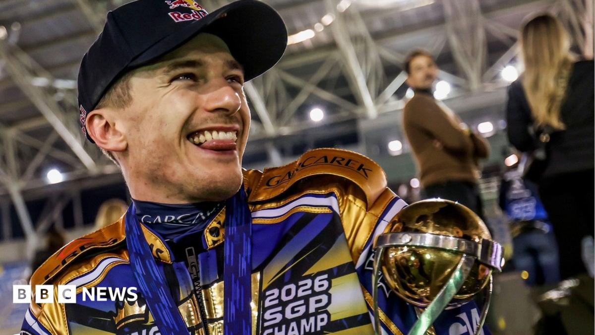 Robert Lambert is grinning and sticking his tongue out. He is wearing a black baseball hat and his black, gold and blue racing outfit. He is cradling his gold trophy.