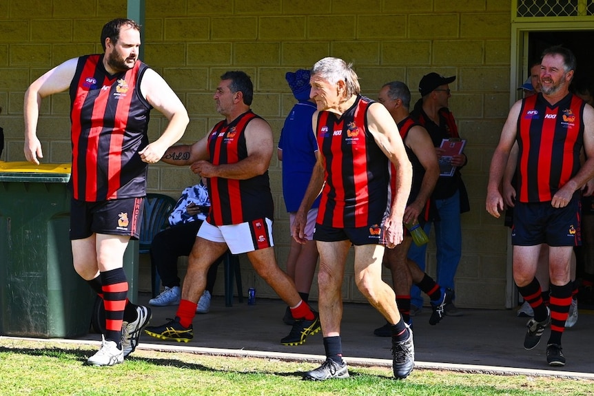 Four men in football guernseys leave the change rooms onto an Australian Rules football oval.