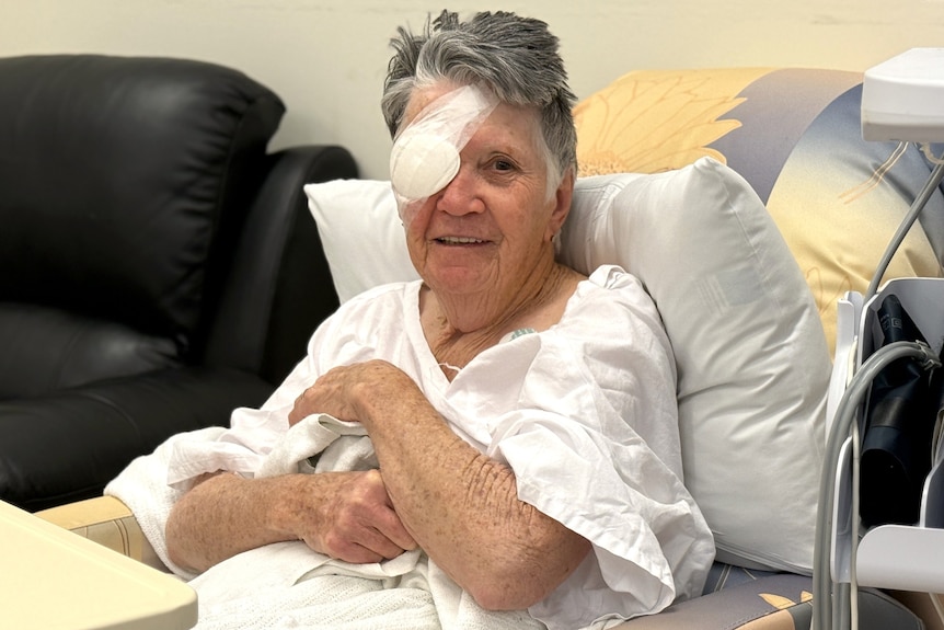 An older woman with short grey hair, sits in a hospital chair with a big bandage over her right eye.