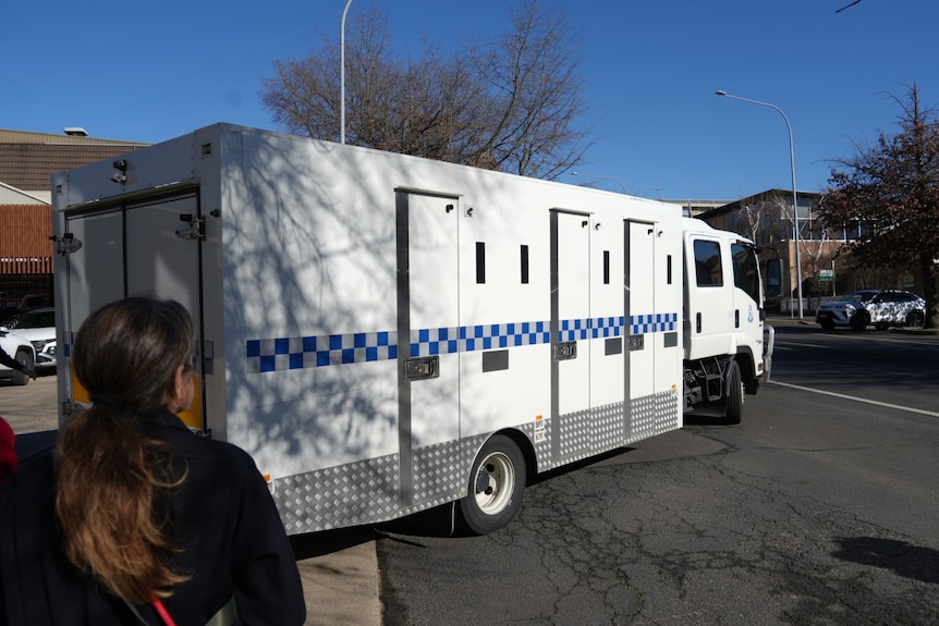 A large white and blue police van backing into a driveway on a sunny day with several people looking on.